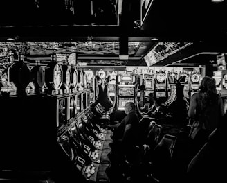 A dimly lit casino filled with rows of slot machines, some occupied by people. The machines' lights contrast with the dark surroundings, creating a vibrant yet subdued atmosphere.