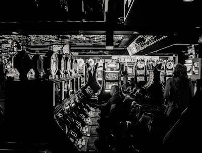 A dimly lit casino filled with rows of slot machines, some occupied by people. The machines' lights contrast with the dark surroundings, creating a vibrant yet subdued atmosphere.