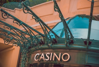 The image displays the entrance to a casino, characterized by detailed and ornate architectural elements with green metal structures and a sign that reads 'CASINO'. The background includes a glass dome and a peach-colored wall.