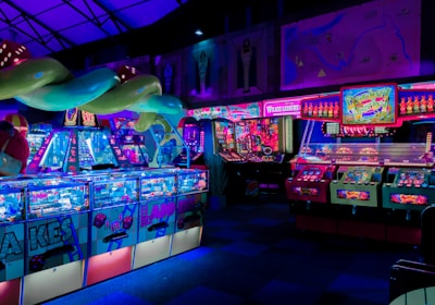 An indoor arcade filled with vibrant gaming machines lit with neon blue and pink lights. The scene includes various arcade games with flashing screens and colorful displays, and a large overhead structure resembling a green snake. The background shows illuminated signs and themed decor, contributing to an immersive entertainment atmosphere.