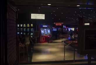 A dimly lit casino with several rows of slot machines, some displaying bright graphics and lights. There are a few people scattered throughout the area, engaging with the machines. The floor is reflective and polished, adding to the ambiance, while the ceiling is dotted with numerous small lights.