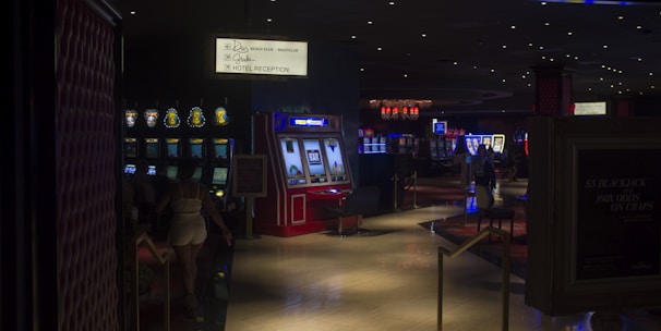 A dimly lit casino with several rows of slot machines, some displaying bright graphics and lights. There are a few people scattered throughout the area, engaging with the machines. The floor is reflective and polished, adding to the ambiance, while the ceiling is dotted with numerous small lights.