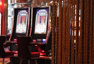 A pair of casino slot machines, each with detailed screens displaying various game outcomes and jackpot amounts. The chairs in front of the slot machines are dark and cushioned for comfort. The background features a hanging curtain made of golden ropes and other casino decorations are visible.