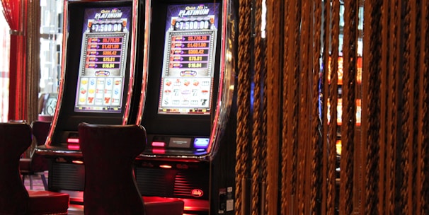 A pair of casino slot machines, each with detailed screens displaying various game outcomes and jackpot amounts. The chairs in front of the slot machines are dark and cushioned for comfort. The background features a hanging curtain made of golden ropes and other casino decorations are visible.