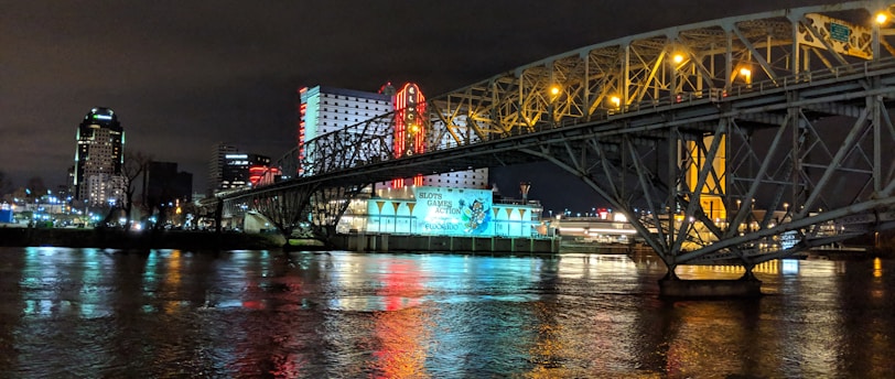 A brightly lit casino with neon signs and red lights is reflected in a river. A bridge stretches across the water, with an urban skyline visible in the background against an overcast night sky.
