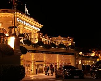 A luxurious casino at night, illuminated with warm, golden lights. The building features elegant architecture with columns and decorative elements. A few people are walking towards the entrance, and there are a couple of parked cars nearby.