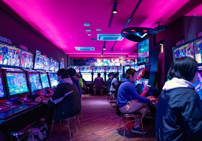 A vibrant arcade with rows of gaming machines adorned with colorful screens. Several people are immersed in playing games, seated on stools in a dimly lit room accented by an intense pink ceiling light. The atmosphere is lively and engaging.