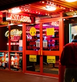 A brightly lit entrance to a casino with neon signs, including one advertising a £500 jackpot. The doors have clear signage indicating entry is restricted to those over 18. A person in a red jacket stands to the right, possibly interacting with a machine or display.