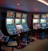 A row of brightly lit slot machines in a casino setting, each with colorful displays and various game themes. The machines are lined up against a dark wall and are unoccupied, with cushioned chairs placed in front of them. The area is dimly lit except for the glow from the screens, creating a vibrant and engaging atmosphere.