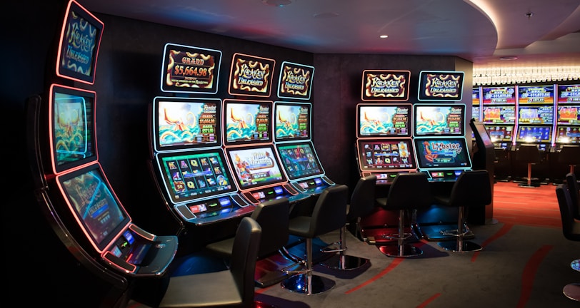 A row of brightly lit slot machines in a casino setting, each with colorful displays and various game themes. The machines are lined up against a dark wall and are unoccupied, with cushioned chairs placed in front of them. The area is dimly lit except for the glow from the screens, creating a vibrant and engaging atmosphere.