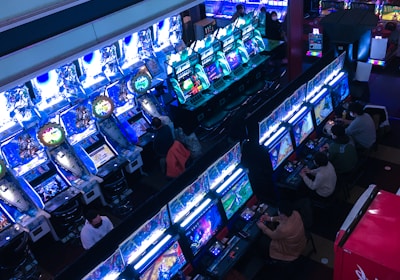 A bustling arcade filled with rows of gaming machines, each with vibrant displays and controls. Several people are seated at these machines, deeply engaged in playing games. Bright fluorescent lights cast a blue and neon glow across the room, enhancing the lively and energetic atmosphere.