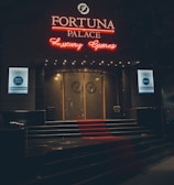 A neon-lit casino entrance with a red carpet leading up to large double doors. The sign reads 'Fortuna Palace Luxury Games' in bright red and white neon lights. The dimly lit atmosphere suggests a night-time setting. There are two digital displays on either side of the entrance showing promotional content.