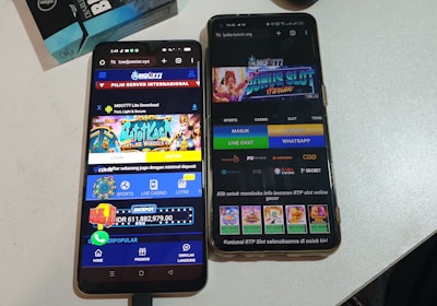 Two smartphones are displayed on a white surface, both showing gambling websites with colorful graphics, text, and various options for logging in and accessing games. The screens are bright and filled with promotional content related to casino and slot games.
